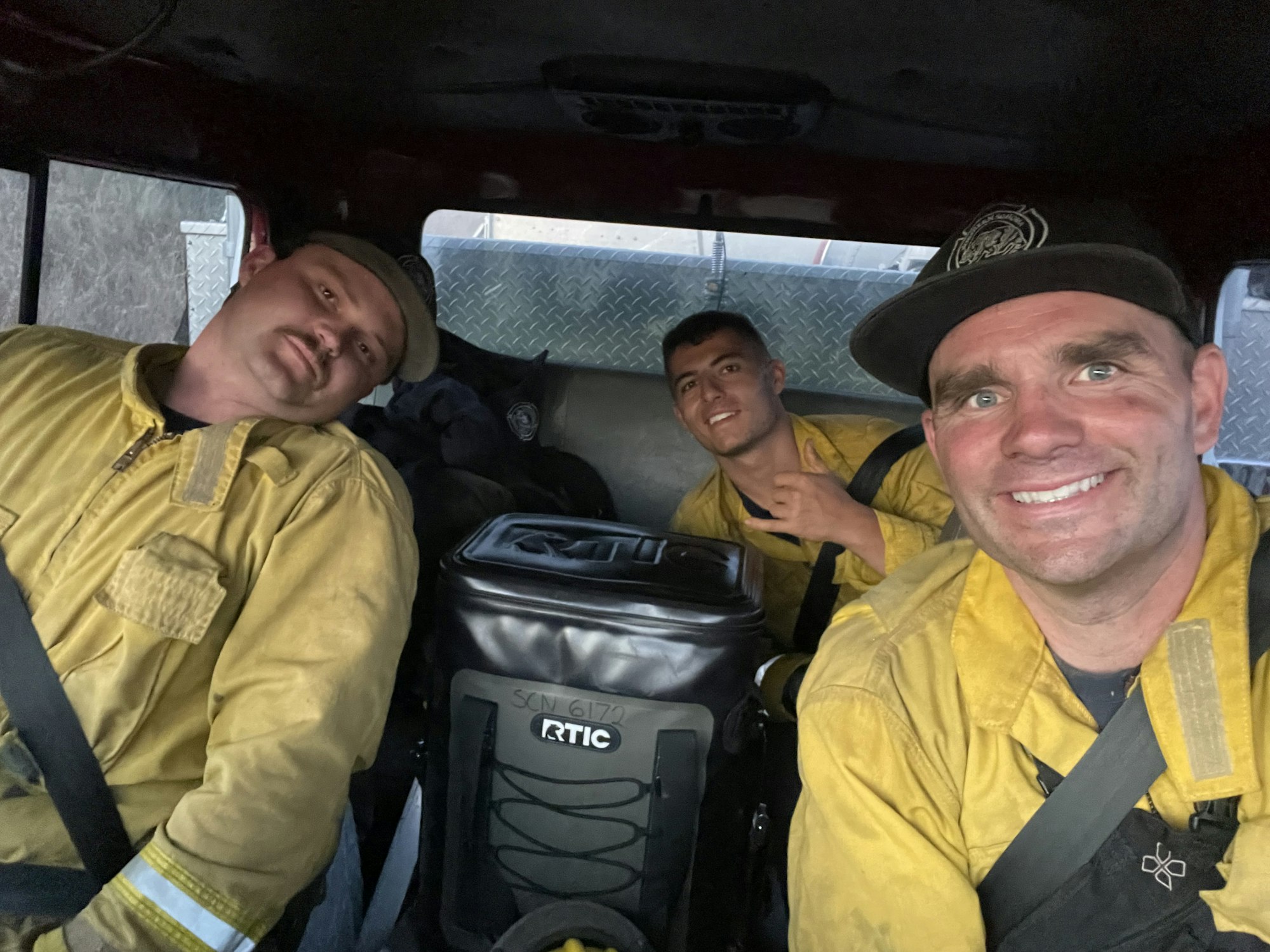 Three smiling firefighters in a vehicle, wearing yellow uniforms, possibly after duty.