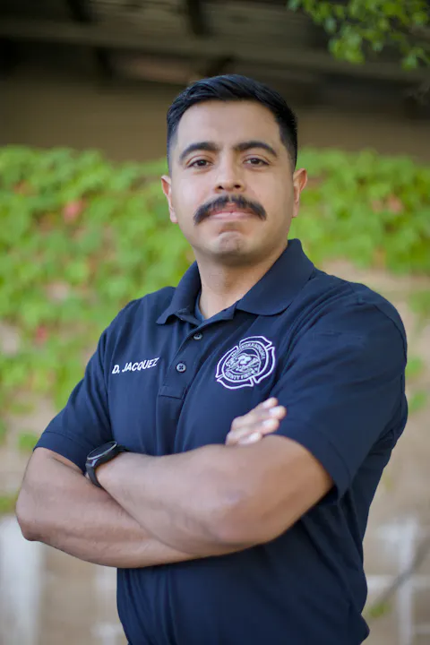 A person in a black polo with a logo and crossed arms stands outdoors.
