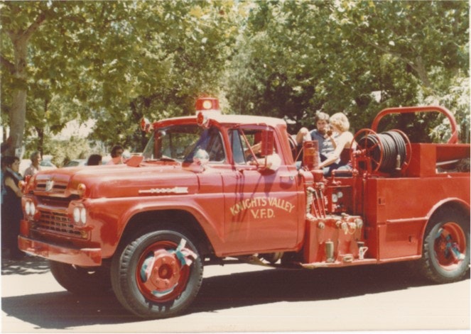 Vintage red fire truck from Knights Valley Volunteer Fire Department in a parade, with people sitting on the back.