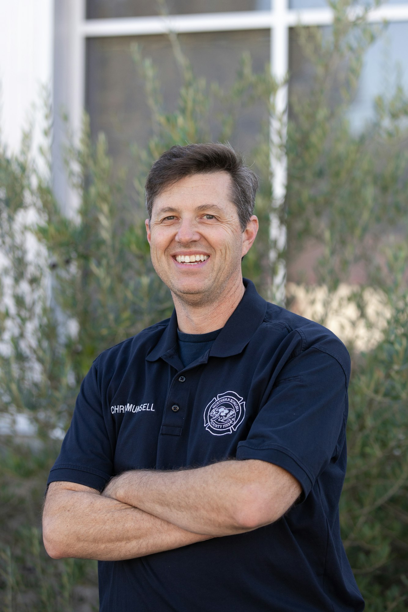 A man is smiling with arms crossed, wearing a dark shirt with a badge, standing outdoors near some greenery.