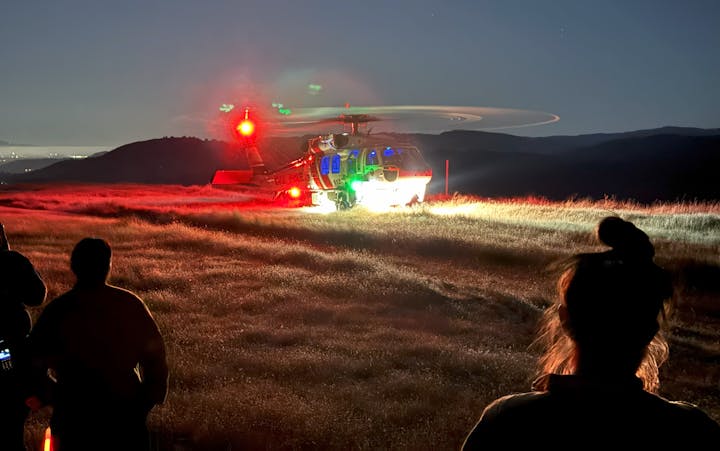 A silhouette of people observing a helicopter landing on a grassy field at dusk with its lights on.
