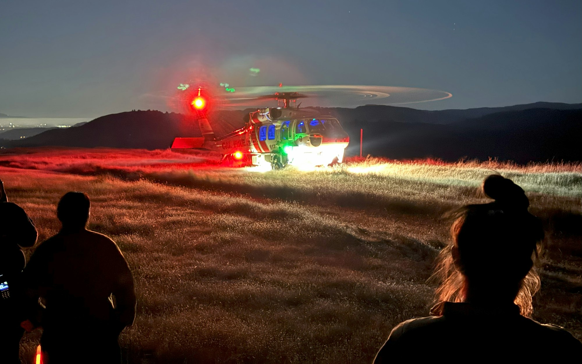 A silhouette of people observing a helicopter landing on a grassy field at dusk with its lights on.