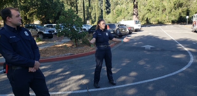 Two firefighters stand in a parking lot; one is gesturing with their arms outstretched.