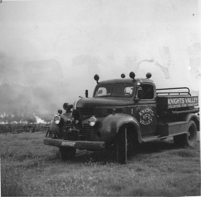 Vintage fire truck labeled "Knights Valley Volunteer Fire Department" on a field next to a fire.