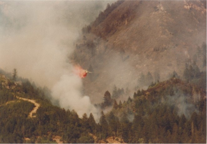 Helicopter dropping water on a forest fire in a mountainous area, with smoke visible.