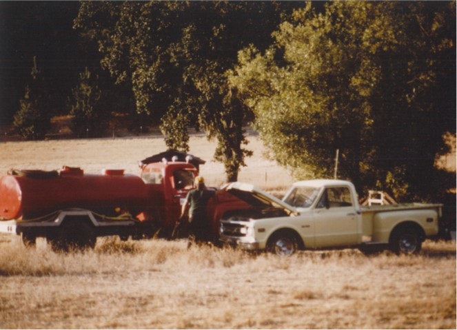 A yellow pickup truck is parked with the hood open next to a large red tanker truck in a grassy area with trees in the background.
