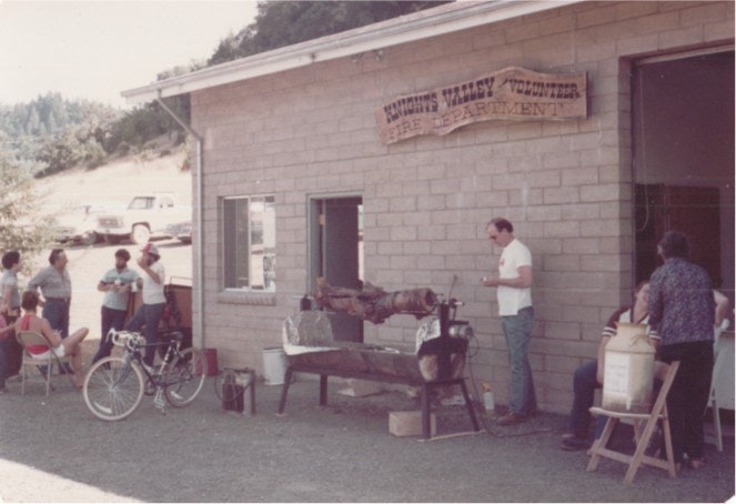 People gathered outside a building with a sign reading "Volunteer Fire Department," a bicycle, and a whole roast on a spit.