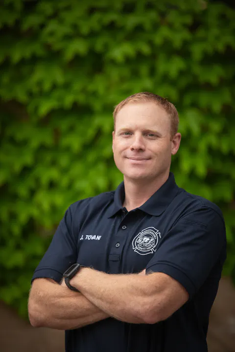A smiling man in a navy polo shirt stands confidently with arms crossed, against a backdrop of green foliage.