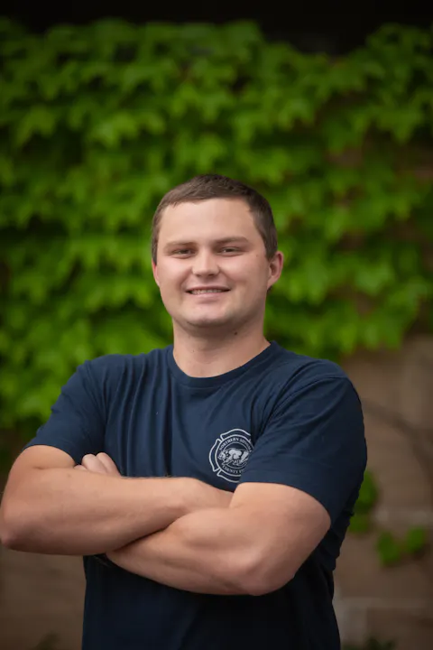 A smiling individual in a dark shirt stands confidently with arms crossed in front of a backdrop of green leaves.