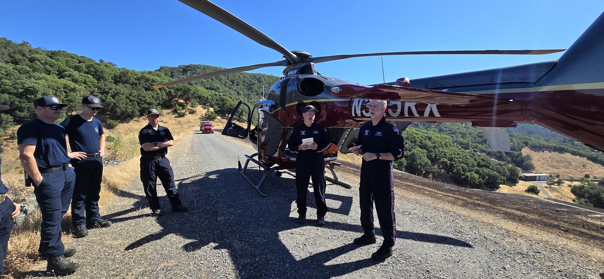 A helicopter and and a group of emergency responders, on a sunny roadside with hills in the background.
