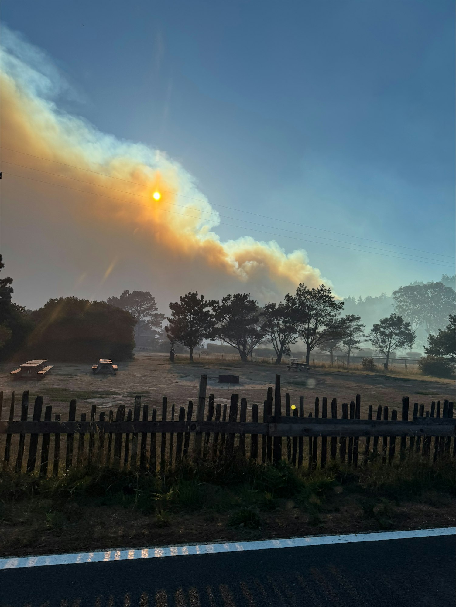 A smoky sky at sunset, with trees and a wooden fence in the foreground, hinting at nearby smoke or fire activity.