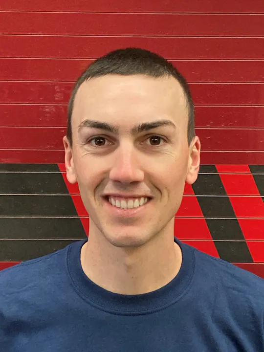 A smiling person with short hair wearing a blue shirt, with a red and black checkered background.