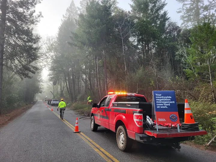 A red utility truck with a sign about vegetation management is parked on a foggy road, with workers and traffic cones visible.