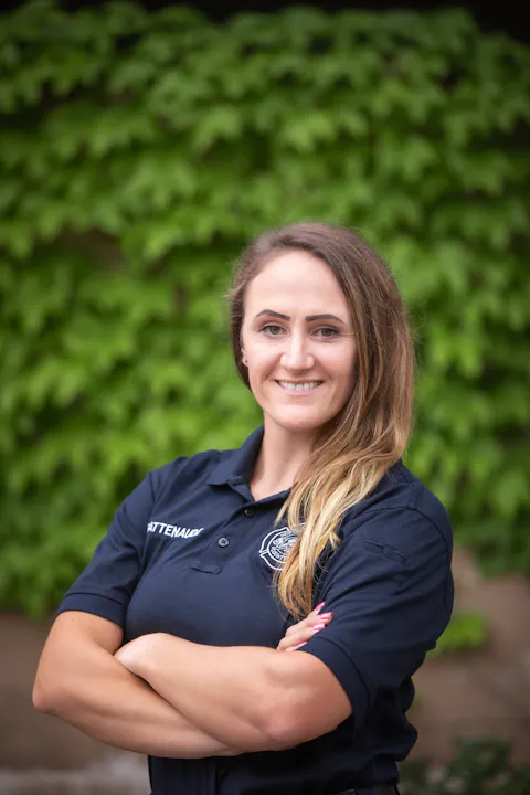A confident person with long hair, wearing a dark polo shirt, stands with arms crossed against a green backdrop.