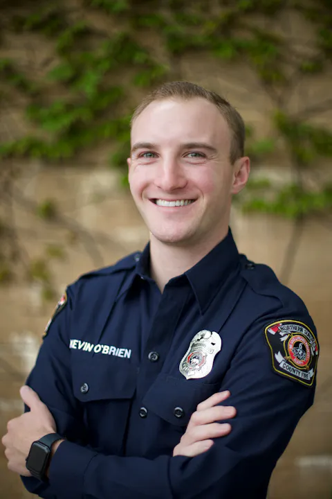 A person in a navy-blue uniform stands smiling with arms crossed, badges visible on the shirt.