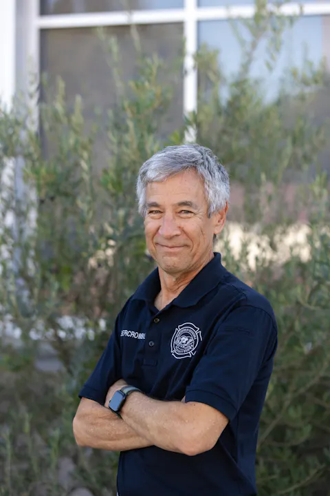 A man with gray hair and a blue shirt stands outdoors, smiling with arms crossed, surrounded by greenery.