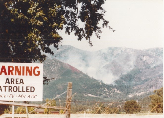 A warning sign with "Area Patrolled" text and mountains in the background emitting smoke.