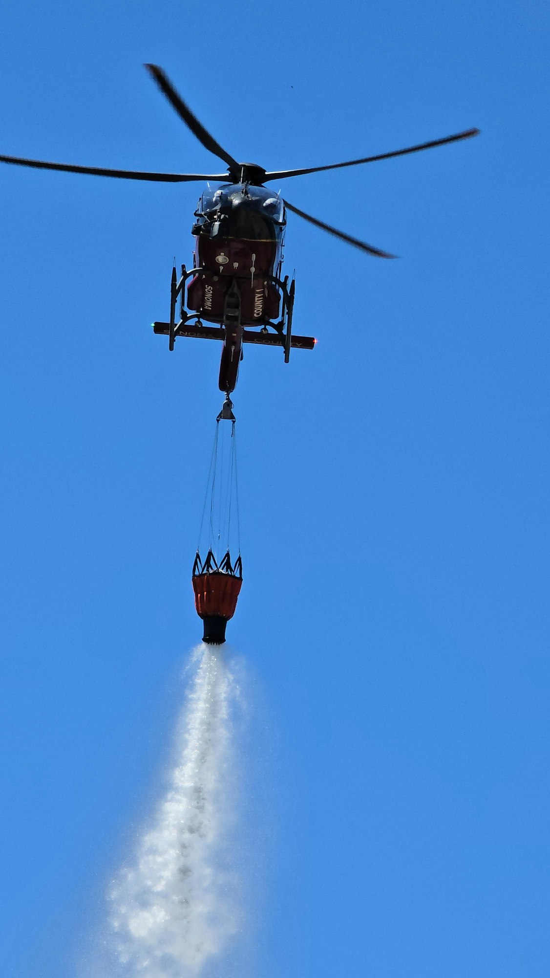 A helicopter with a water bucket is dispersing water mid-flight against a clear blue sky.