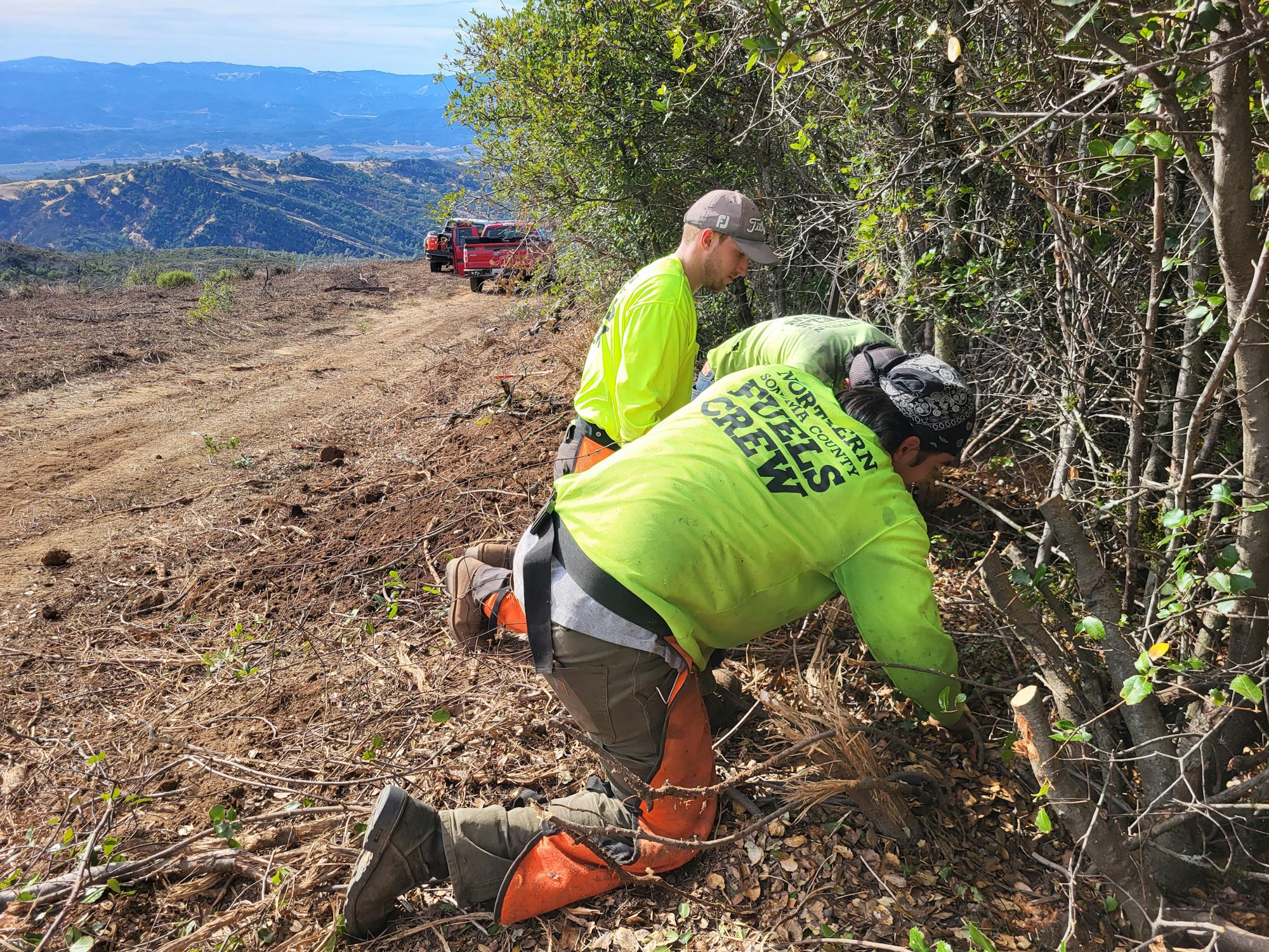 Two workers in bright shirts are clearing brush on a hillside with trucks in the background.