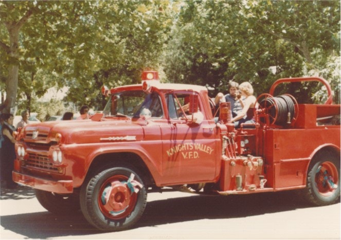 A vintage red fire truck with "Knights Valley V.F.D." on the side, carrying people, in a sunny outdoor setting.