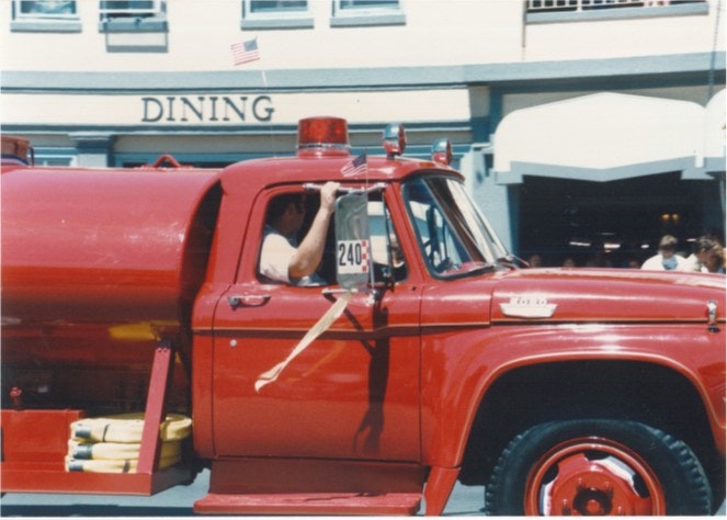 Red fire truck in a parade with flags, passing a building labeled "DINING".