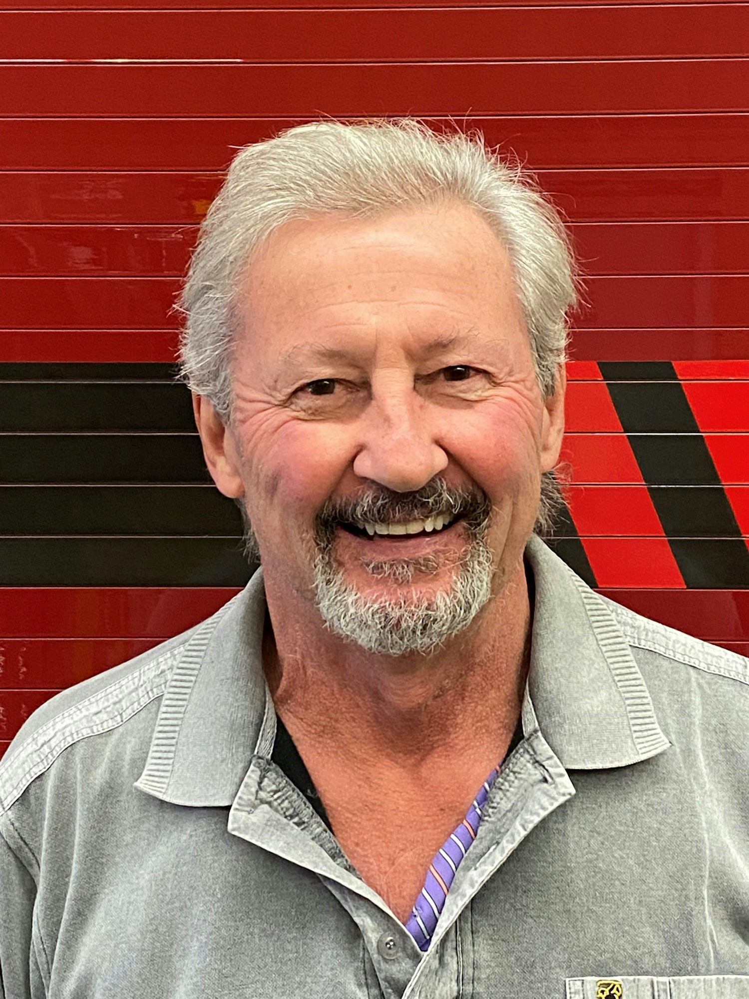 A smiling man with a mustache and white hair against a red background with black and red patterns.
