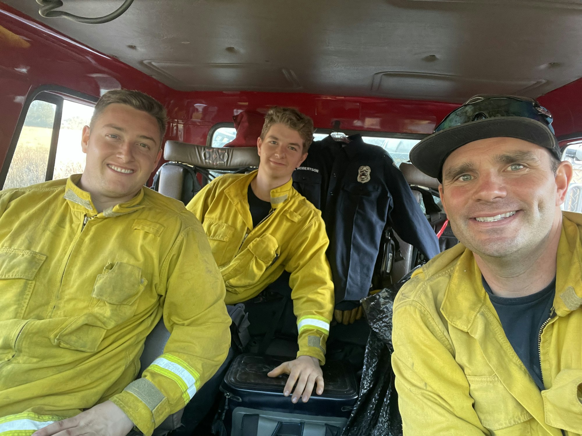 Three firefighters in yellow uniforms sitting inside a red vehicle, smiling at the camera.