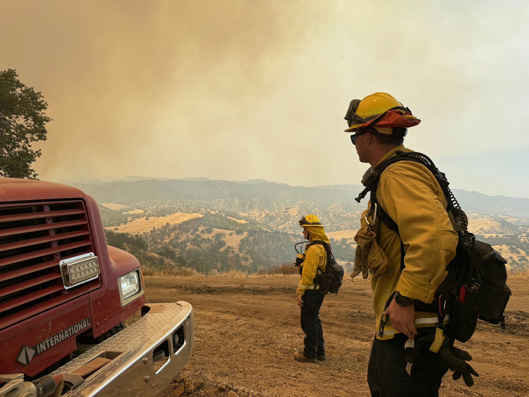 Two firefighters in yellow gear near a fire truck, overlooking a smoky landscape.