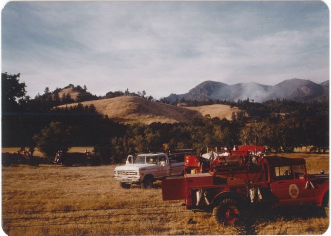 Fire trucks in a grassy field with rolling hills and mountains in the background.