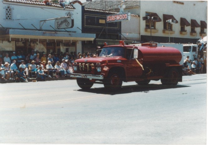 A vintage red tanker truck parades past a crowd seated on the sidewalk in front of shops.