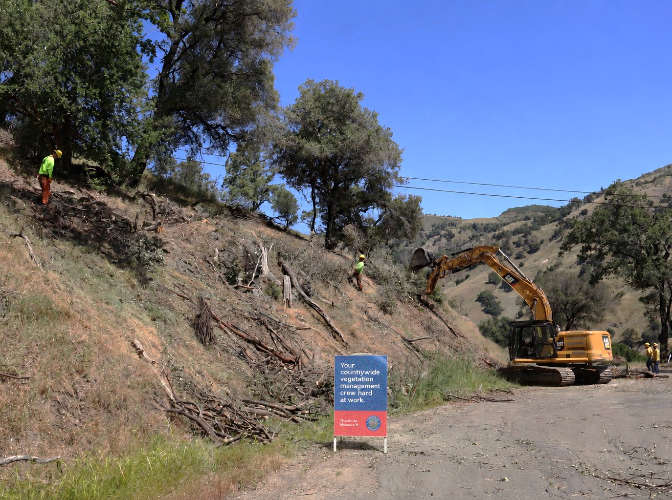 Workers are managing vegetation along a hillside, using machinery while a sign acknowledges their efforts. Clear blue sky above.