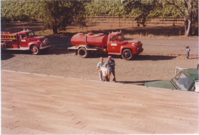 Two vintage red fire trucks parked near trees, with three people standing nearby on a gravel path adjacent to a shingled roof.