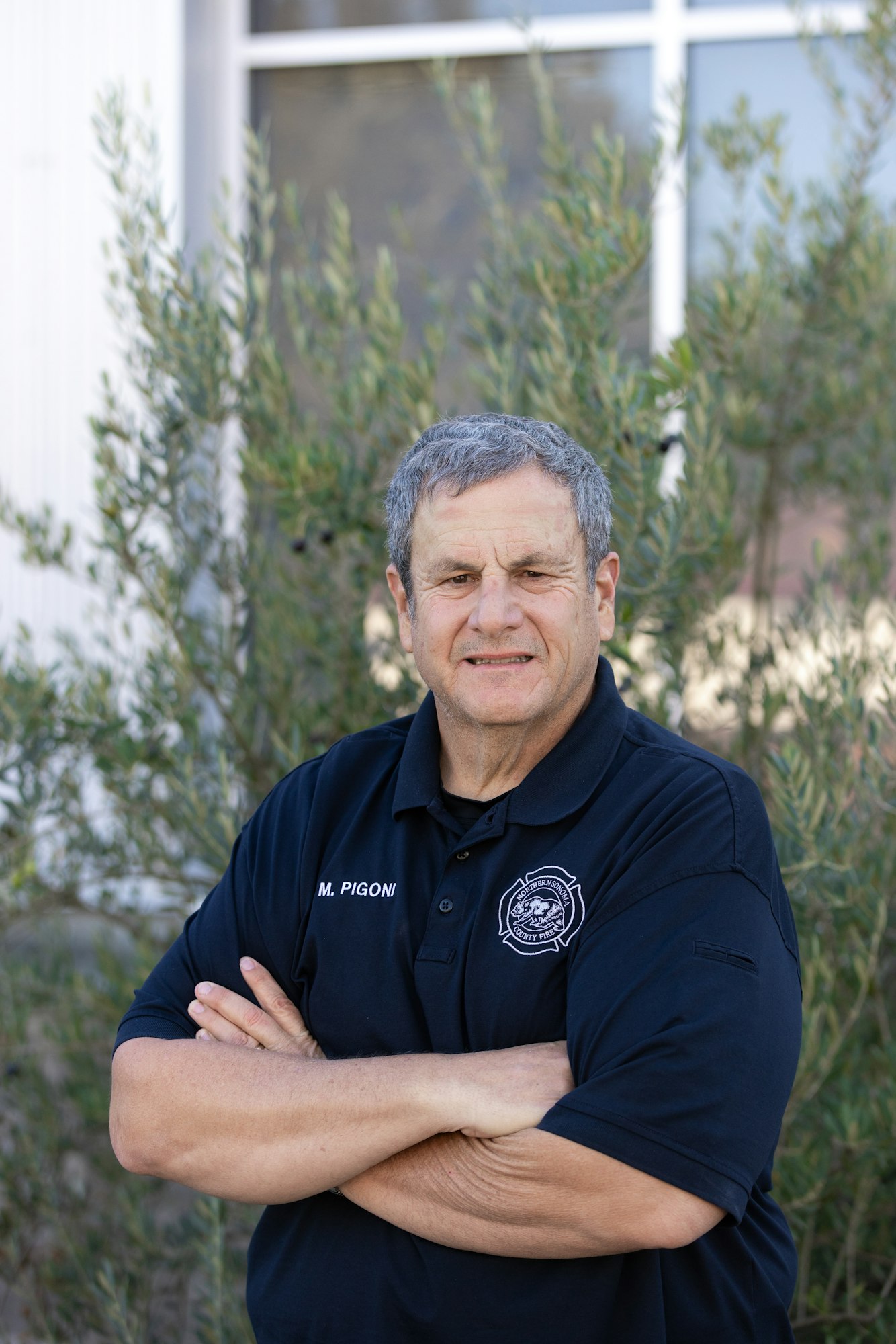 The image features a person with crossed arms, wearing a black polo shirt with a badge, standing outdoors near greenery.