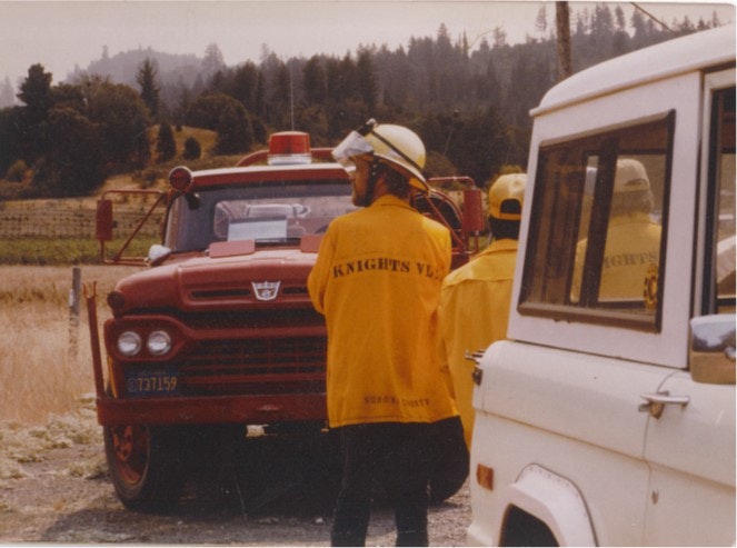 A fire truck and firefighters in yellow jackets, with "Knights Valley" visible, stand in a rural area near a white vehicle.