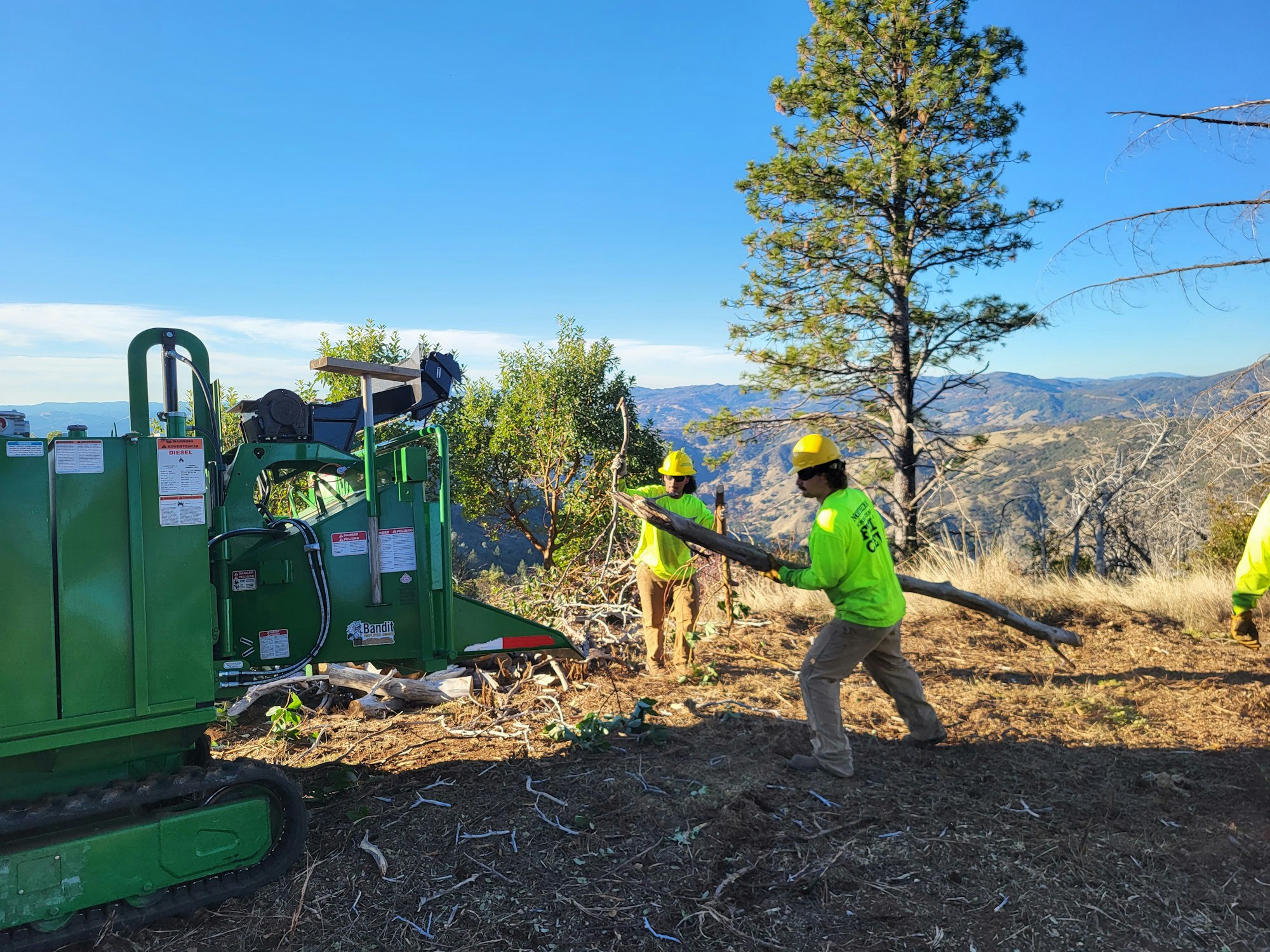 Workers in green shirts use a wood chipper on a hillside with scenic views.