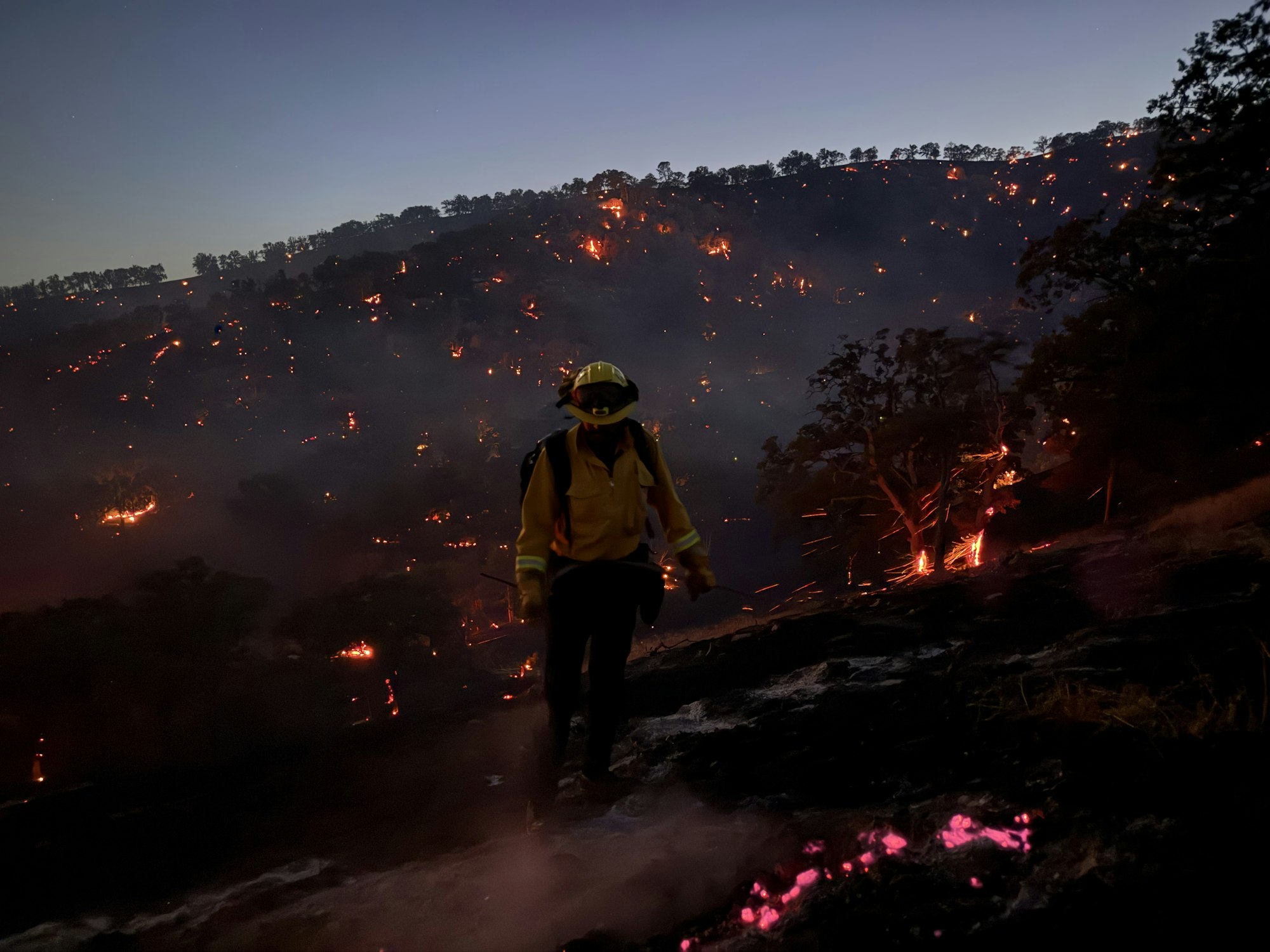 A firefighter stands near a smoldering hillside at dusk, with small flames visible.