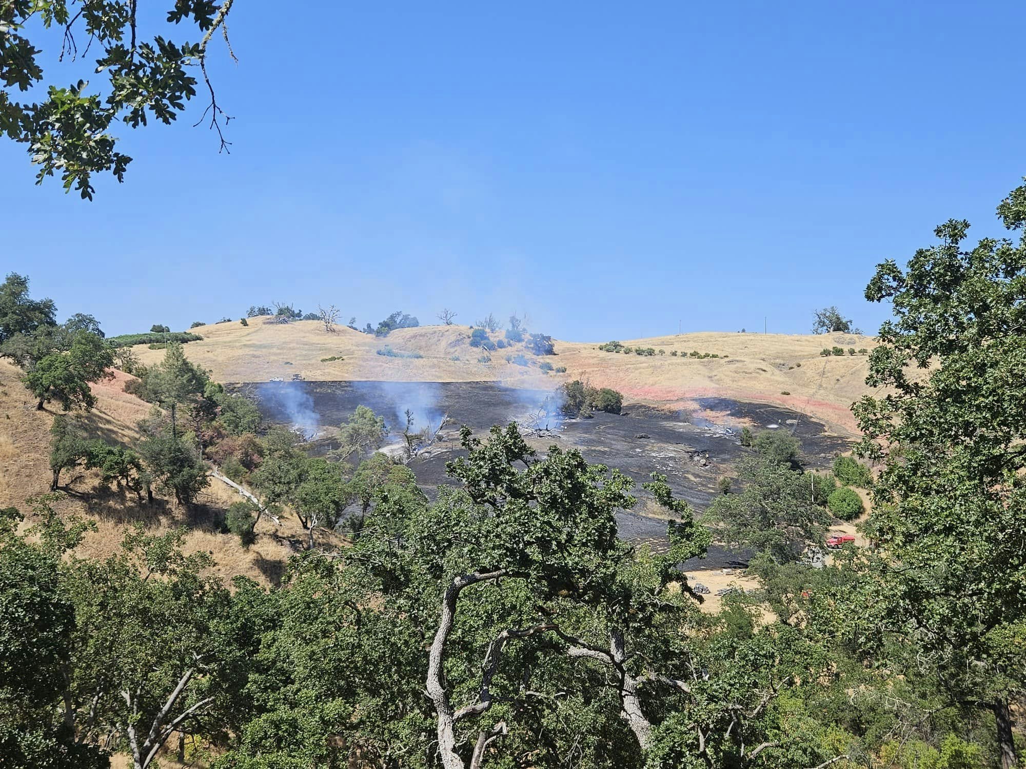 A dry landscape with smoke rising from burned areas, surrounded by trees and clear blue skies, indicating recent fire activity.