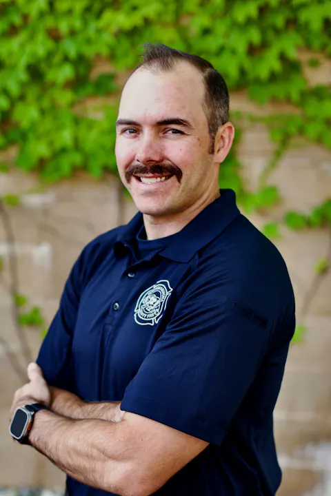 A person with a mustache, wearing a dark blue shirt with an emblem, stands against a green leafy background, smiling with arms crossed.