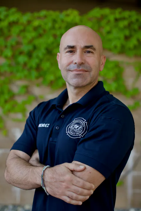 A person in a dark blue shirt with a fire department logo stands with arms crossed against a leafy background.