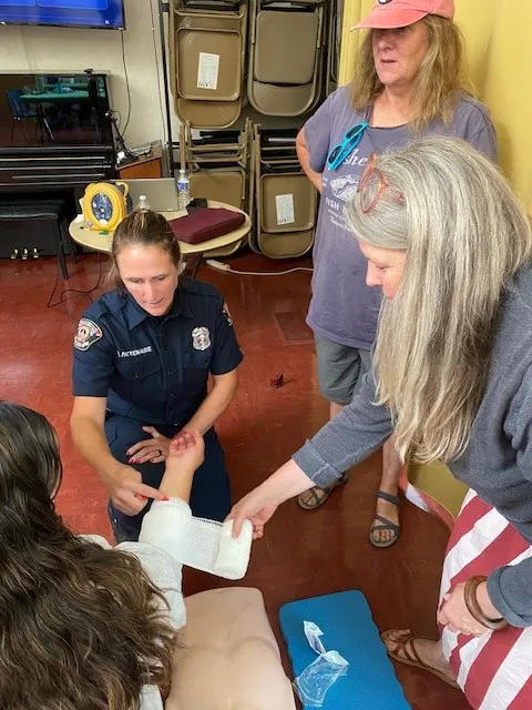 A paramedic tends to a person with a bandaged arm, while two women observe, in a casual indoor setting.