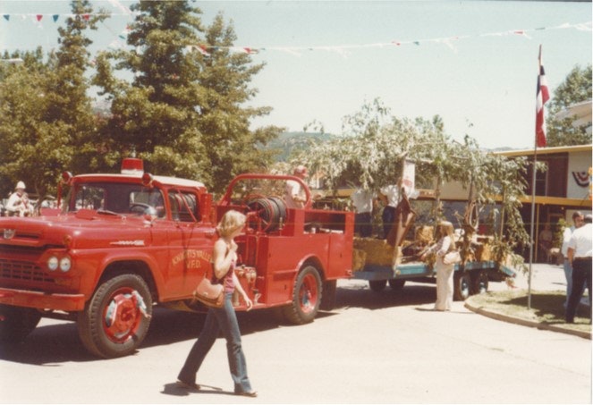 A red fire truck in a parade, pulling a trailer with trees, people nearby, and festive decorations overhead.