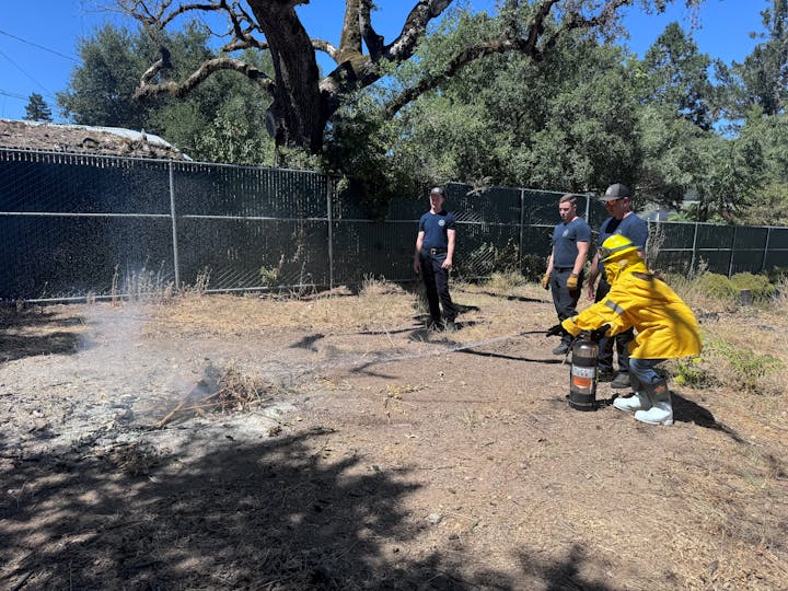 Female in nomex training with an extinguisher on a small fire, observed by three others outdoors near trees and a fence.