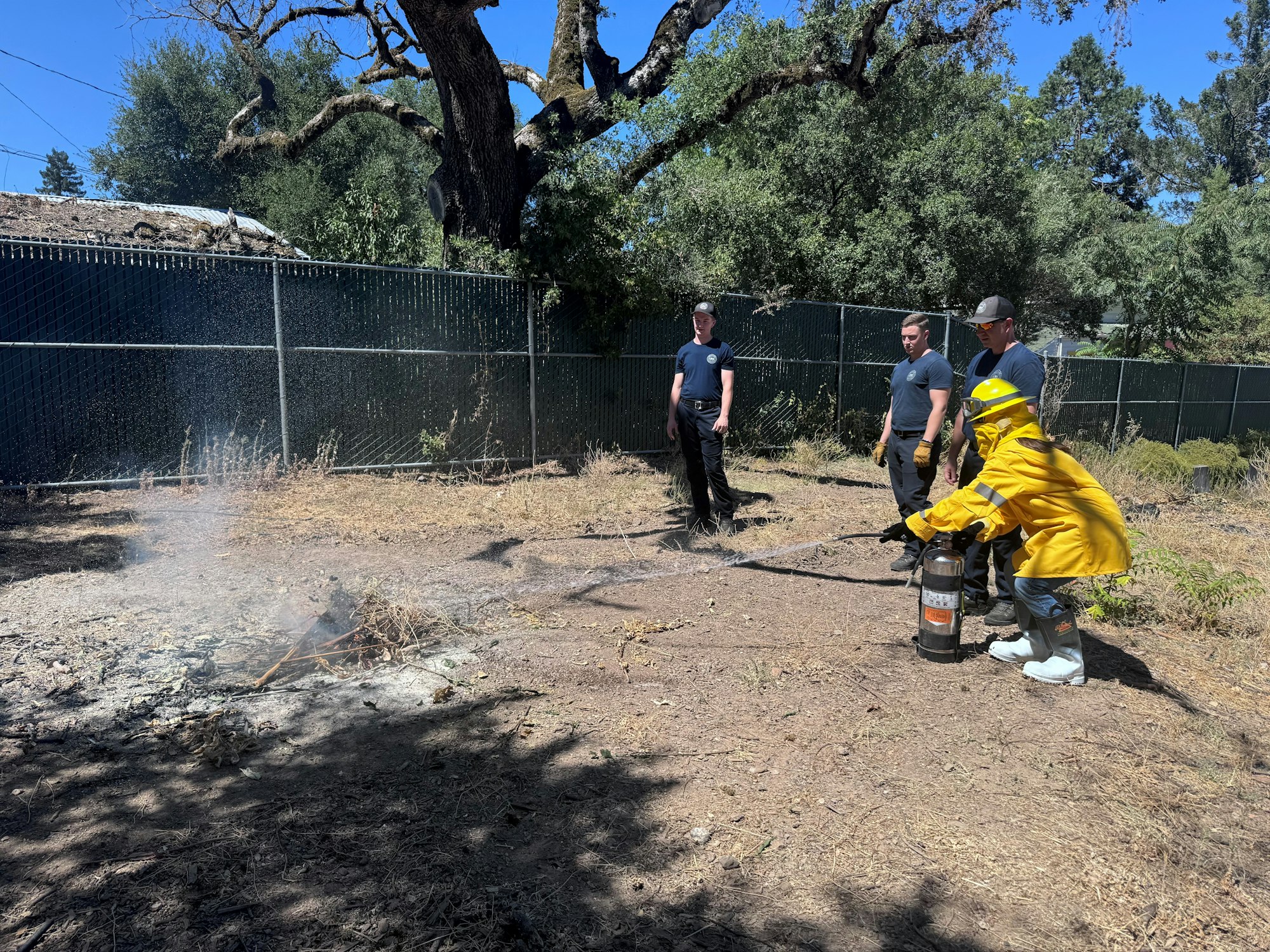 Female in nomex training with an extinguisher on a small fire, observed by three others outdoors near trees and a fence.