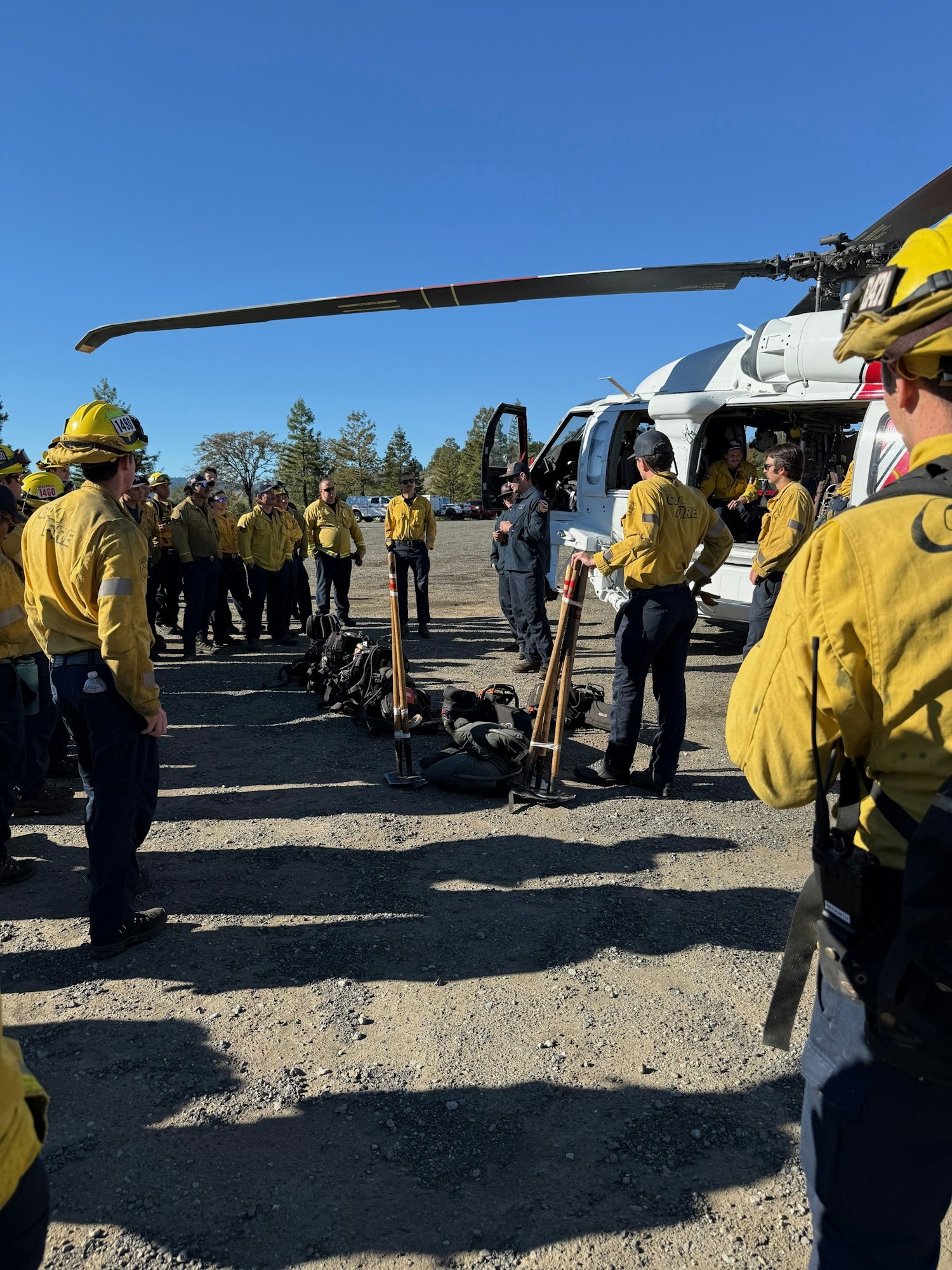 Firefighters in yellow uniforms gather for a briefing near a helicopter, preparing for a response operation.