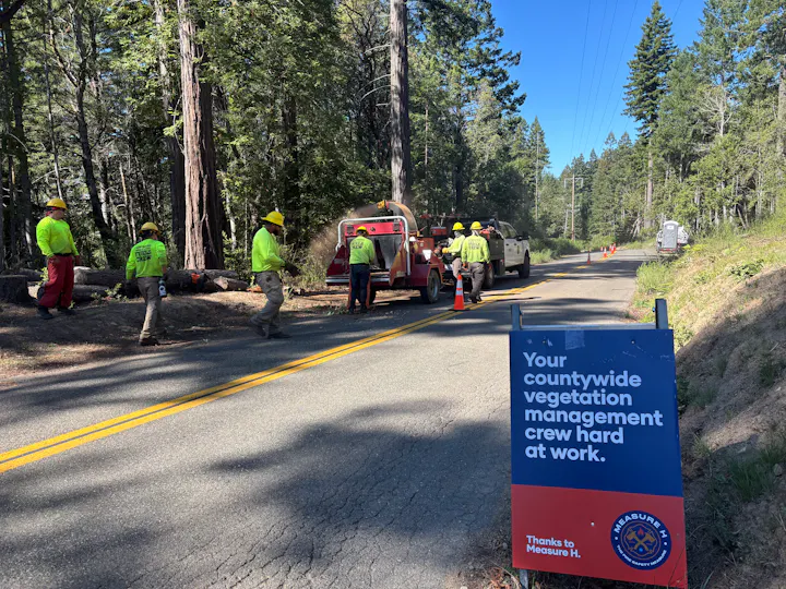 Workers in high visibility yellow feeding chipper alongside road with Measure H a-frame