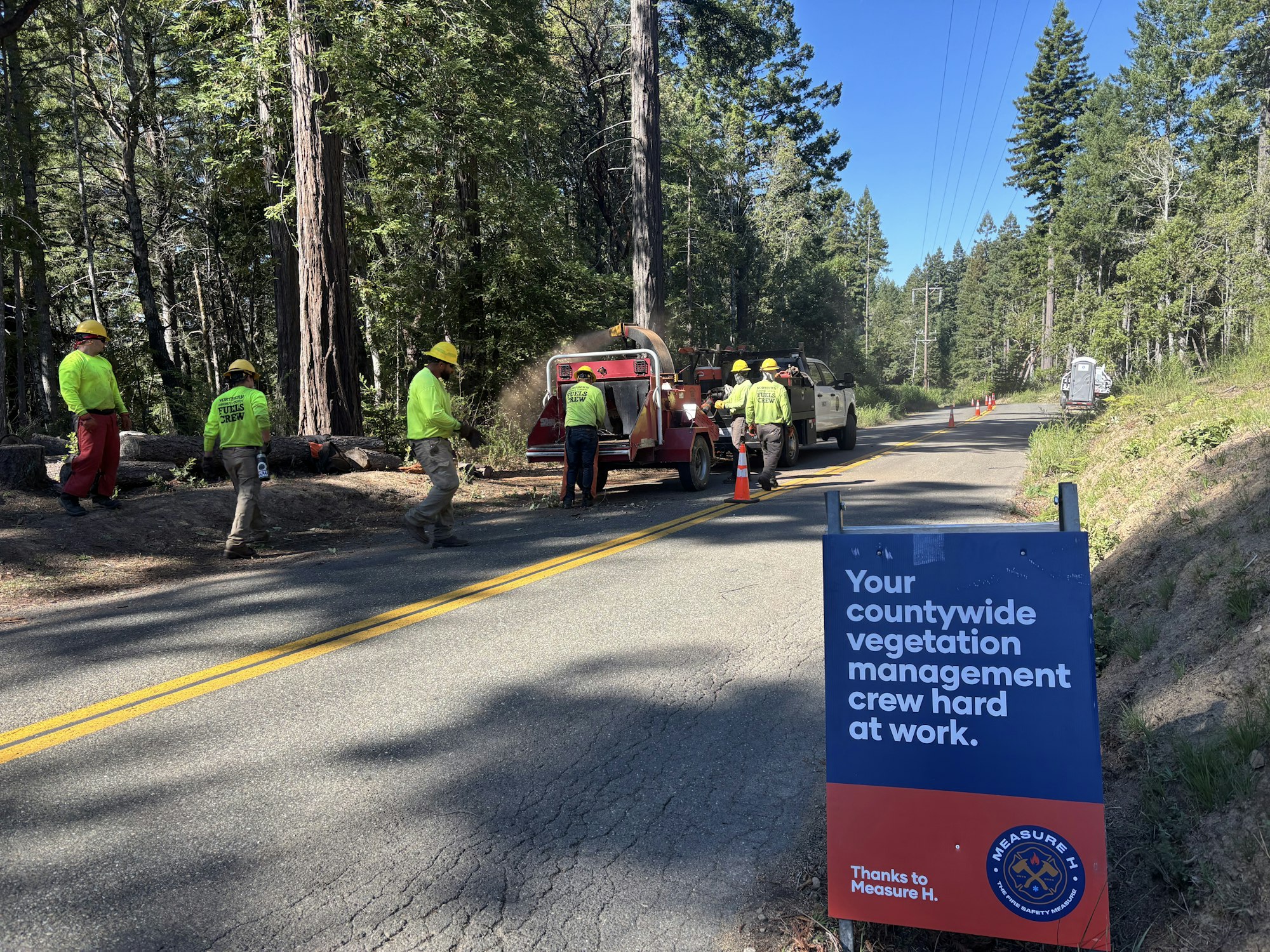 Workers in high visibility yellow feeding chipper alongside road with Measure H a-frame