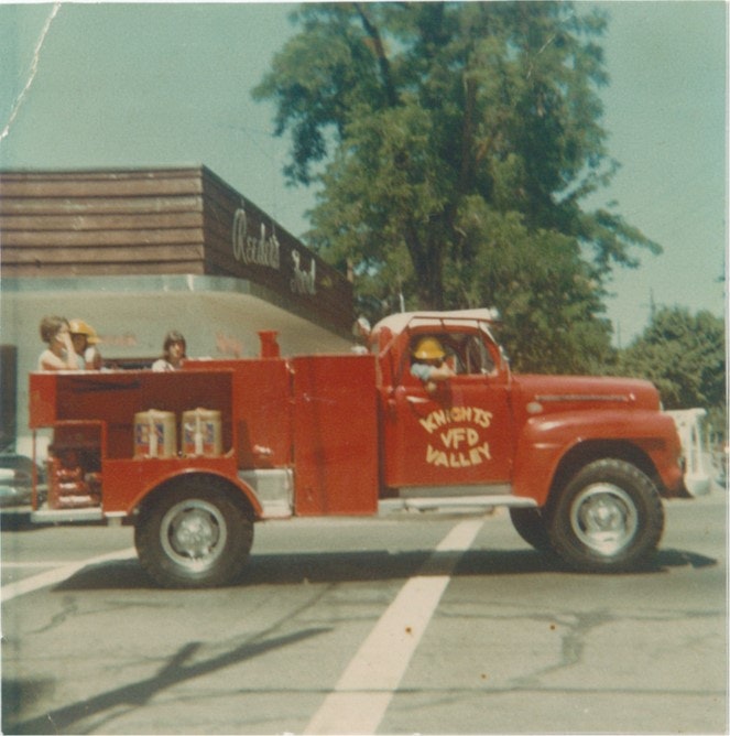 A vintage red fire truck labeled "Knights VFD Valley" with people in yellow helmets, parked near a building with trees in the background.