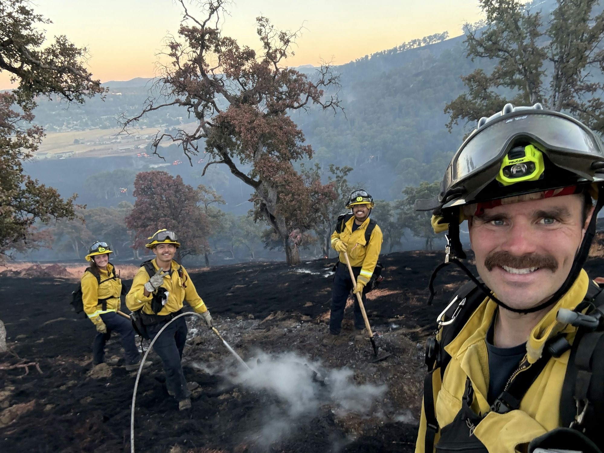Firefighters in yellow uniforms work on a charred landscape, smiling, with burnt trees around them.