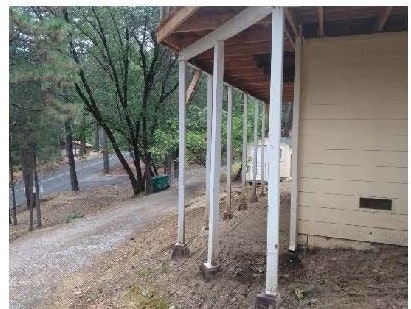 House on a slope with elevated wooden supports, dirt ground, trees, and a driveway or path.