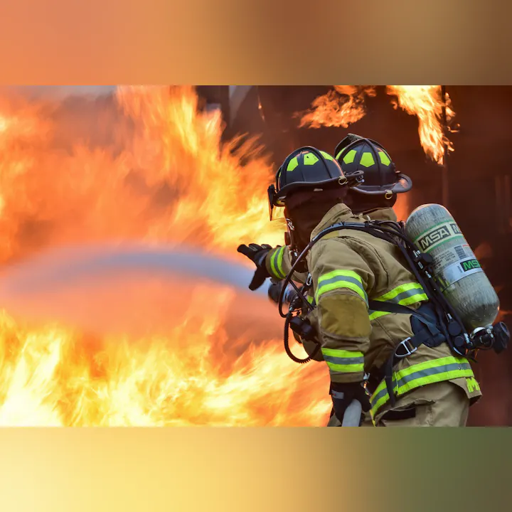 Two firefighters in protective gear combat a large blaze, using a hose to spray water on the flames.